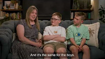 A woman and 2 children are sitting on a sofa, talking to each other. There's a closed caption that reads 'And it's the same for the boys'.