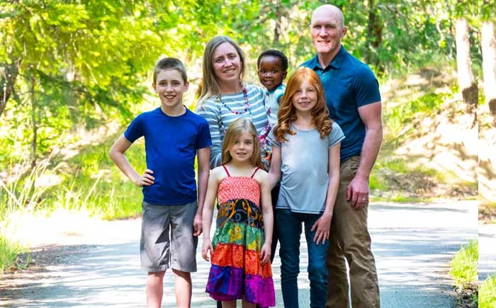 A family of two adults and four children standing outside in a woodland area, smiling at the camera.